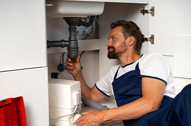 Ein Handwerker in blauer Arbeitskleidung liegt auf dem Boden und führt eine Reparatur unter einem Waschbecken durch. Neben ihm steht ein offener Werkzeugkasten, und auf dem Boden liegen verschiedene Rohrteile.